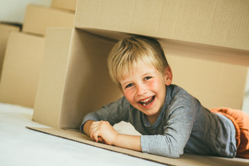 Rose cheeks preschooler boy sitting inside box in new house. Child lying on the floor, laughing and looking froom cardboard box. Toned.