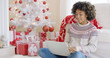 © Dash - Young woman typing on a laptop at Christmas sitting on the floor in front of the tree and a pile of colorful red and white gifts.
