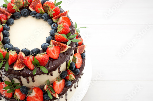 Zdjęcie XXL Dwuwarstwowy tort w czekoladzie, ozdobiony plasterkami truskawek, jagód, fig i zielonych liści na białym drewnianym stole. Obraz menu lub katalogu słodyczy. Widok z góry.