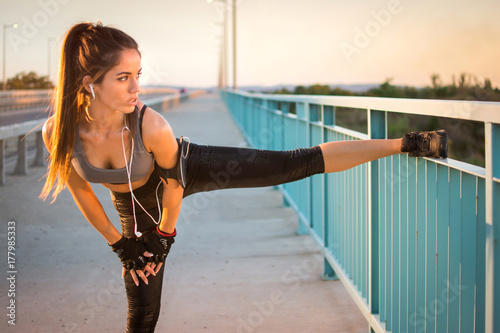 Plakat Sportowa dziewczyna w sportswear rozciąga jej nogi nad mosta ogrodzeniem outdoors.