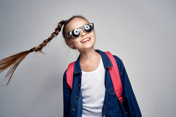 Wall Mural - stylish schoolgirl smiling with glasses on shoulders backpack on gray background