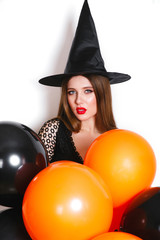 Portrait of beautiful young woman in black witch halloween costume with orange and black balloons over white background