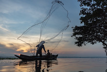 Asian Fisherman On Wooden Boat Casting A Net For Catching Freshwater Fish In Nature River In The Early Morning Before Sunrise.