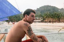 Young Man With Tatooed Arm And No Shirt, Sitting On A Sailboat On The Beach Of A Tropical Island