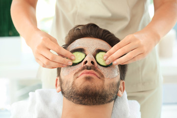 Canvas Print - Beautician putting fresh cucumber slices on young man's face in spa salon