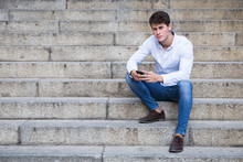 Handsome Young Man In Shirt And Jeans Sitting On Stairs And Using His Smartphone Looking At Camera