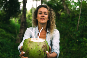 Wall Mural - Young woman with curly hair is offering a coconut cocktail on a green jungle background. Green coconut with straws in hands of a tourist female walking the tropical forest during summer vacation.