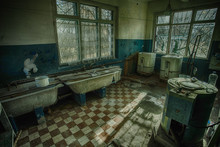 Sinister And Creepy Old Laundry Room With A Dirty Floor And Broken Wash Machines And Bathes In An Abandoned Psychiatric Hospital.