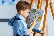 Cute, Serious And Focused, Three Years Old Boy In Blue Shirt And Jeans Apron Painting On Canvas Standing On The Easel. Concept Of Early Childhood Education, Talent, Happy Family Or Parenting