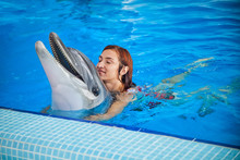 A Cheerful Red-haired Woman In A Red Bathing Suit Laughs, Bathes And Holds A Dolphin In The Blue Pool Behind Her Back