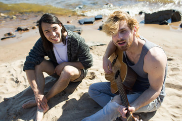 Wall Mural - Young man playing guitar and singing on seashore with his friend on background