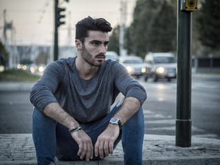 Wall Mural - Attractive young man portrait at night with city lights behind him in Turin, Italy, sitting on sidewalk curb