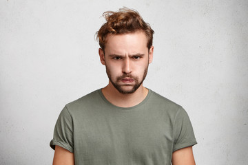 Wall Mural - Portrait of sad bearded young man has grumpy expression pouts his lips after quarrel with wife. Discontent male student looks gloomy after failing exam at university. Negative human emotions.