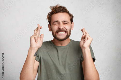 Cuadros en Lienzo Cheerful bearded man keeps fingers crossed, smiles broadly and closes eyes, makes desire wish