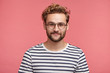 © wayhome.studio - Indoor shot of pleasant looking bearded hipster guy wears spectacles and striped t shirt, looks directly into camera, isolated over pink studio background. Male student spends weekends at home alone