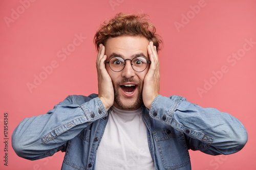 Fotografia Indoor shot of amazed excited man doesn`t believe his success, keeps hands on head, stares at camera, says omg or wow