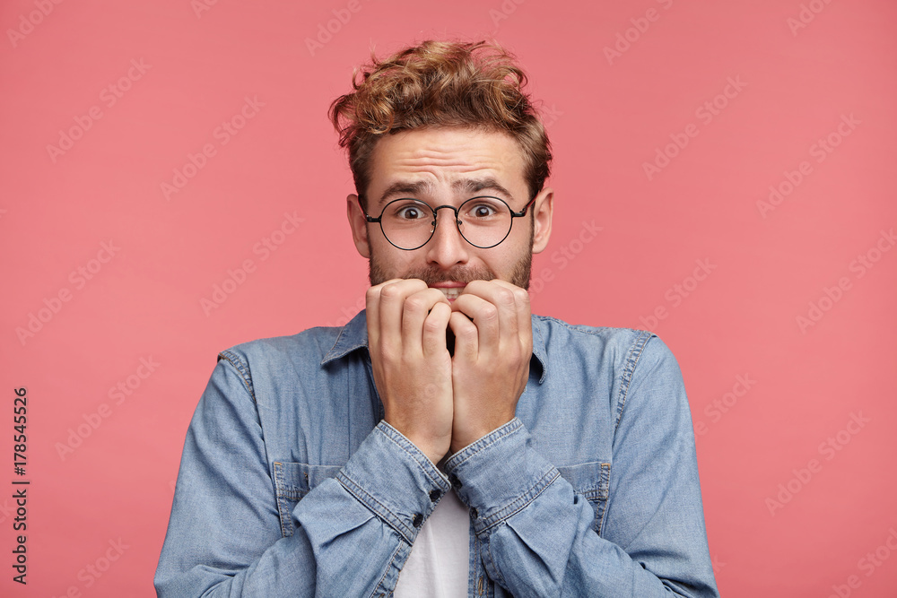 Worried hipster guy bites nails, looks nervous before passing exam or important event in his life. Embarrassed fashionable young man being afraid of difficulties, stands against pink background - obrazy, fototapety, plakaty 