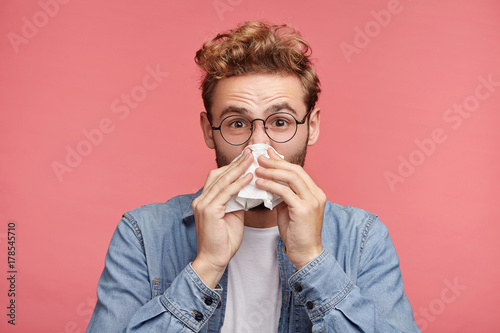 Fotografía Indoor shot of bearded man has running nose, rubs nose with handkerchief, being ill, caught cold after long walk outdoors on rainy weather