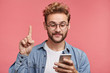 © WHstudio Leushin N - Young curly stylish hipster guy dressed in jean shirt, holds smart phone, reads something attentively, raises index finger, quotes someone words, proves his rightness. People and technology concept