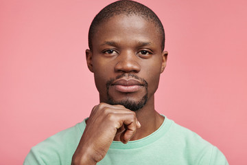 Wall Mural - Horizontal portrait of confident serious attractive dark skinned man keeps hand under chin, has full lips, wears casual t shirt, isolated over pink background. Thoughtful male manager poses in studio