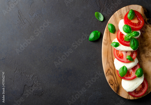 Fotografía Sliced tomatoes and mozzarella on a wooden board made of olive tree, dark stone background