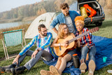 Fototapeta Londyn - Multi ethnic group of friends dressed casually having fun playing guitar during the outdoor recreation with tent, car and hiking equipment near the lake