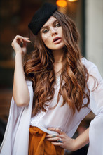 Outdoor Waist Up Portrait Of Young Fashionable Woman Wearing Hat With Veil Posing In Street. Model Has Beautiful Long Hair. Lady Wearing Stylish Clothes. Lights On Background. Female Fashion Concept