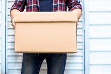 Young Man Holding A Moving Cardboard Box In Front Of A Storage Door.Life Style, Storage, Moving, Storing, Organizing Concept. Space To Write