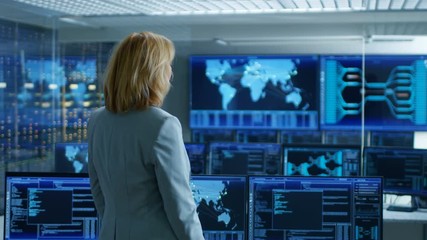 Fototapete - In the System Control Room Female Head of the Department Crosses Arms and Thinks Standing with Her Back to the Camera. In the Background Multiple Screens Showing Interactive Data. 