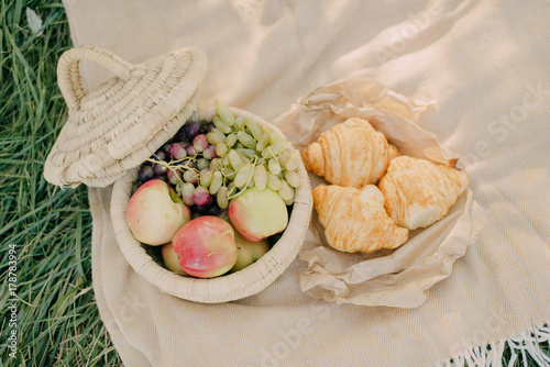 Plakat na polanie wiklinowy kosz z winogronami i jabłkami obok rogalików