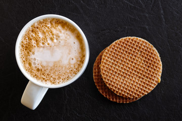 Wall Mural - Milky frothy coffee in white mug next to a couple of round waffle biscuits isolated on black leather from above.