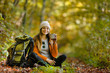 © proimagecontent - Beautiful brunette tourist girl wears cap and backpacked have break with cup of tea sitting in the forest, autumn tourism concept