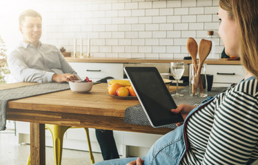 Young married couple in the kitchen. A man sits at a table and with a smile looks at his wife. A pregnant woman sits at a table and uses a tablet computer. Social network, lifestyle. Online education.