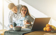 © foxyburrow - Front view.Morning, breakfast.Young married couple in kitchen.Pregnant woman is sitting at table and using laptop, man is holding her pregnant belly, looking on computer monitor. Lifestyle.