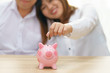 © petzshadow - Smile couple putting a coin into a pink piggy bank on wooden desk - save money for the future.