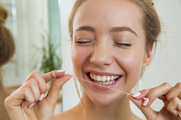 Wall Mural - Young beautiful woman cleaning her teeth with floss in bathroom. Standing in towel, looking in the mirror, laughing and having fun. Daily routine. Beautiful smile with white teeth.