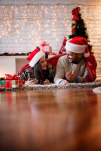 Romanic Happy Couple With Santa Hats Lying On A Carpet And Playing Under A Red Blanket At Warm Home For Christmas Holidays.