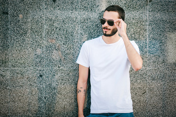 Wall Mural - Young man wearing white blank t-shirt with beard in glasses, standing on the street on city background. Street photo