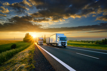 Wall Mural - small truck overtakes heavy truck on the asphalt road in rural landscape at sunset with dark clouds