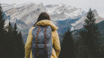 Young Caucasian female hiker in yellow raincoat wearing backpack enjoys the mountain view in French Alps