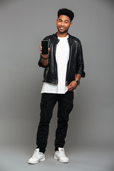 Poster - Full length photo of smiling fashionable african guy standing with hand in his pocket while showing blank mobile screen, looking at camera