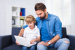 © Di Studio - computer and internet concept - young father and son sitting on sofa with laptop