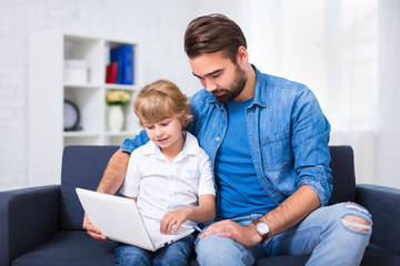 Wall Mural - computer and internet concept - young father and son sitting on sofa with laptop