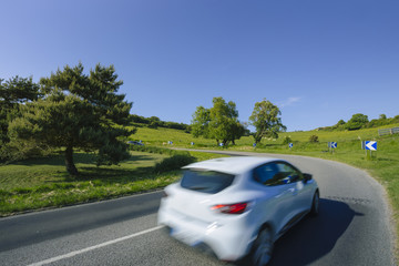 Car driving on the asphalt curvy road through green fields and forests on a sunny day in Normandy, France. Countryside landscape, sunbeams in the blue sky, road network and transportation concept