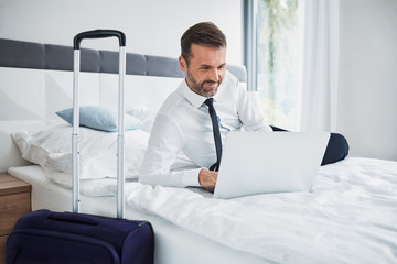 Sticker - Happy businessman using laptop and lying on bed while in hotel room on business trip