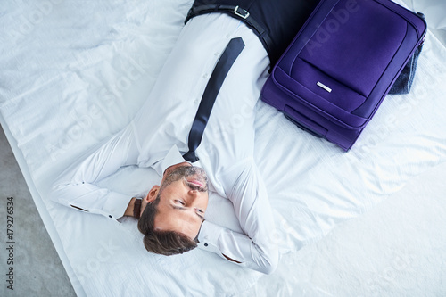 Plakat Zmęczony biznesmena lying on the beach na łóżku w pokoju hotelowym po przyjeżdżać na podróży służbowej