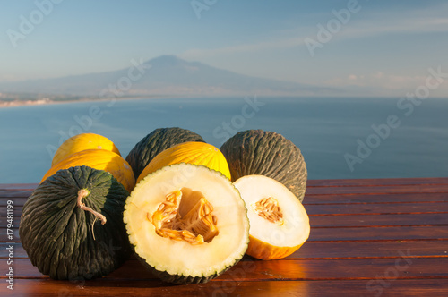 Cuadros en Lienzo Typical mediterranean fruits: yellow melon with blue sea and Mount etna in the b