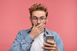 © WHstudio Leushin N - Horizontal portrait of shocked terrified stylish man covers mouth with hand, looks at smart phone as reads horrified news, isolated over pink background. Facial expressions, technology concept