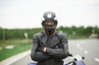 Lifestyle portrait of self determined confident young blue eyed male motorcyclist dressed in stylish leather outfit and helmet, standing outdoors next to his motorbike, keeping arms crossed