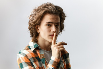 Wall Mural - Don't say anybody. Studio shot of cute guy with wavy hair having mournful expression, holding index finger at his lips, asking to keep his secret. Human emotions, feelings, reaction and body language